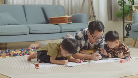 full body of asian father and sons lying on the floor in the room with plastic toy brick. looking at smartphone, drawing, talking, playing together at home