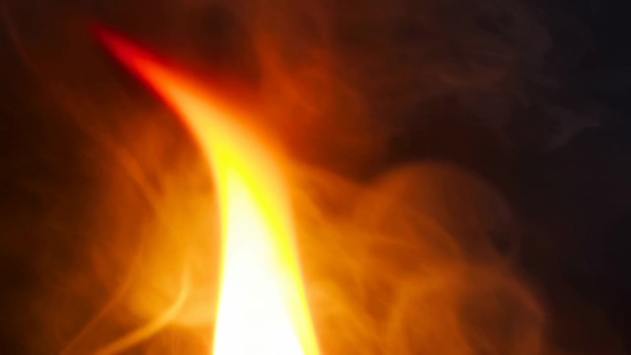 Tightly cropped view of narrow flame rising through dark space with yellow white luminous core, orange body, red tapered tip, and soft smoke swirling around glowing column, with flame, fire, glow