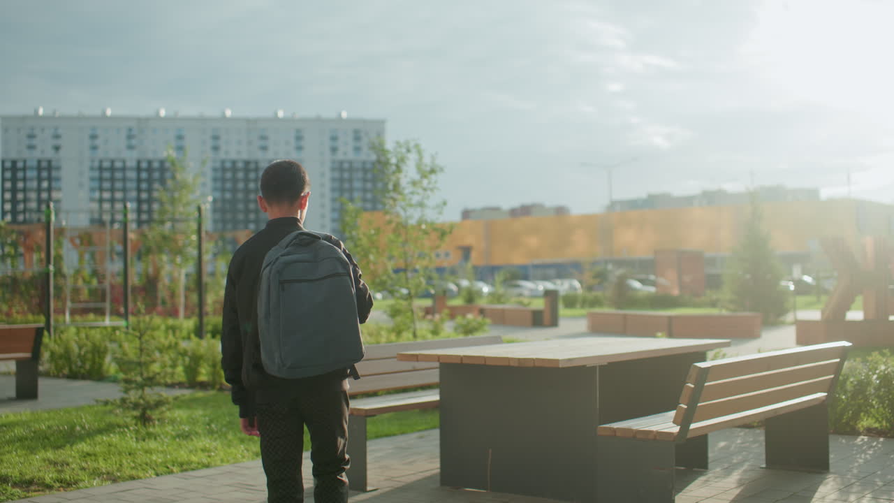 Rear view of boy walking outdoors with backpack toward wooden bench and table in urban park, sunlight casting warm glow over scene with blurred modern buildings in background and greenery around