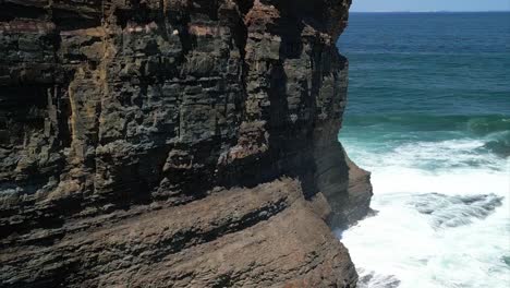 Closeup-drone-shot-of-bands-of-iron-in-the-cliffs-spinning-around-the-point-to-reveal-caves-being-carved-out-by-the-ocean