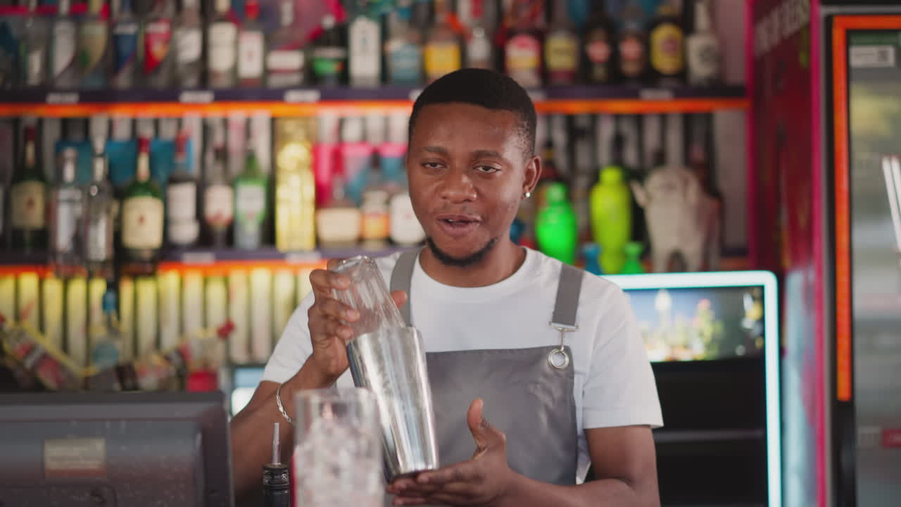 Barman shakes drink in shaker at counter in nightclub. Young black bearded bartender prepares party drinks for guests. African American man enjoys work