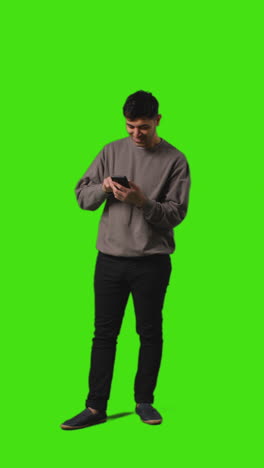 Vertical-Video-Full-Length-Shot-Of-Man-Looking-At-Text-Message-On-Mobile-Phone-Standing-Against-Green-Screen-3