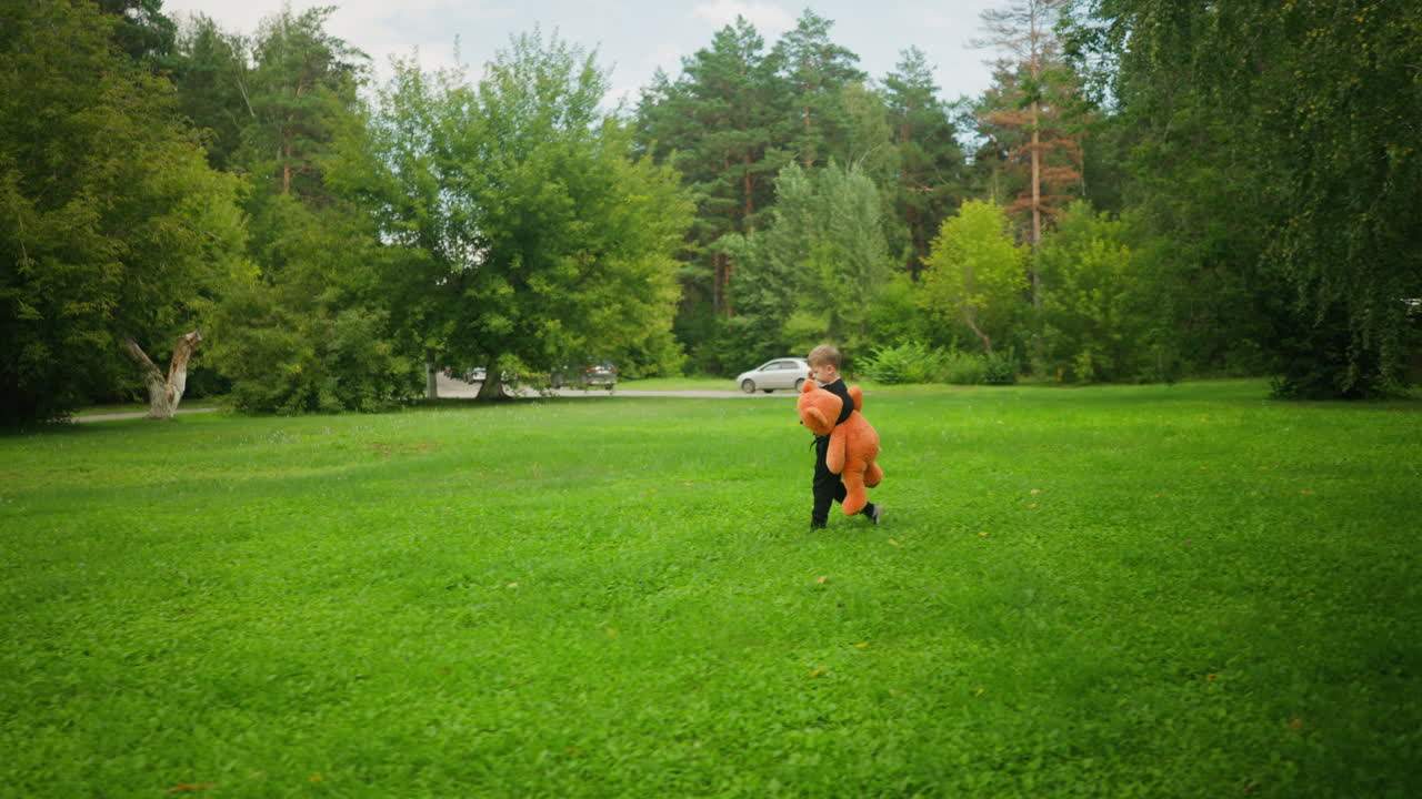 Little boy dressed in black walking alone across open grassy field carrying large teddy bear with moving vehicle in background surrounded by lush trees and distant parked cars under bright sky