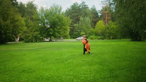 little boy dressed in black walking alone across open grassy field carrying large teddy bear with moving vehicle in background surrounded by lush trees and distant parked cars under bright sky