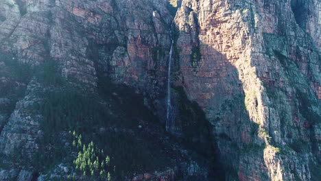 Aerial---Ascending-drone-reveals-beautiful-waterfall-dropping-from-sheer-cliff-of-imposing-sandstone-mountain