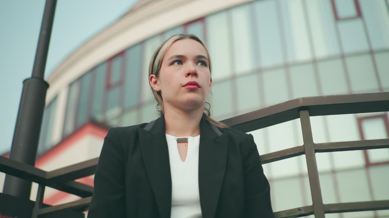 Close up of bored woman in professional outfit looking tired with iron railing behind her and glass building in background under clear sky