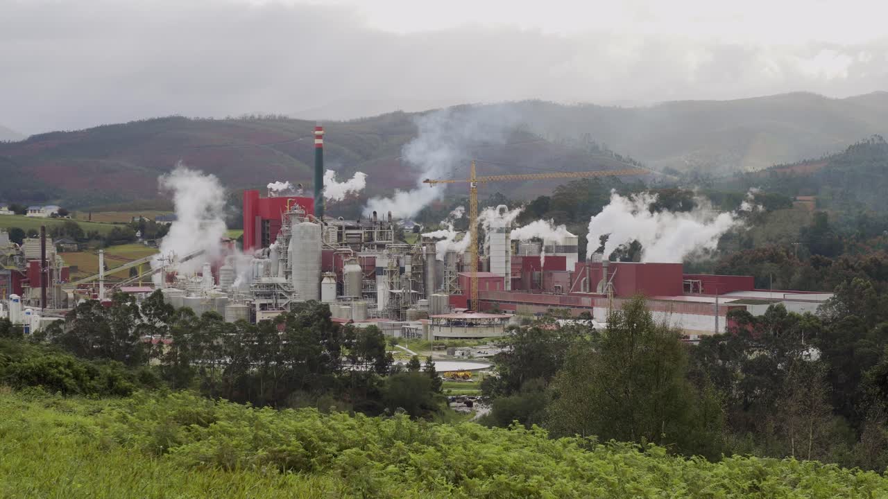 una industria del papel que emite humo y gases contaminantes de muchas chimeneas en un área rural verde mientras el viento dispersa la contaminación en un día nublado