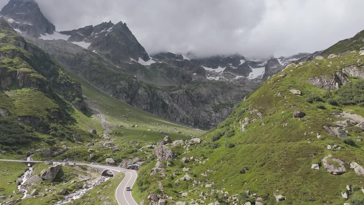 Yazın Interlaken'e doğru giden yemyeşil bir İsviçre Alpleri vadisi üzerinde drone uçuşu; virajlı dağ yolu, kayalık yamaçlar ve alçak bulutlar altındaki dramatik zirveler.