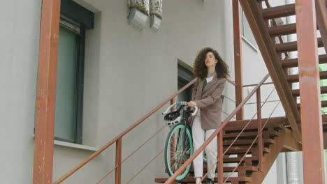 Curly Caucasian woman in formal clothes carrying her bike down the stairs