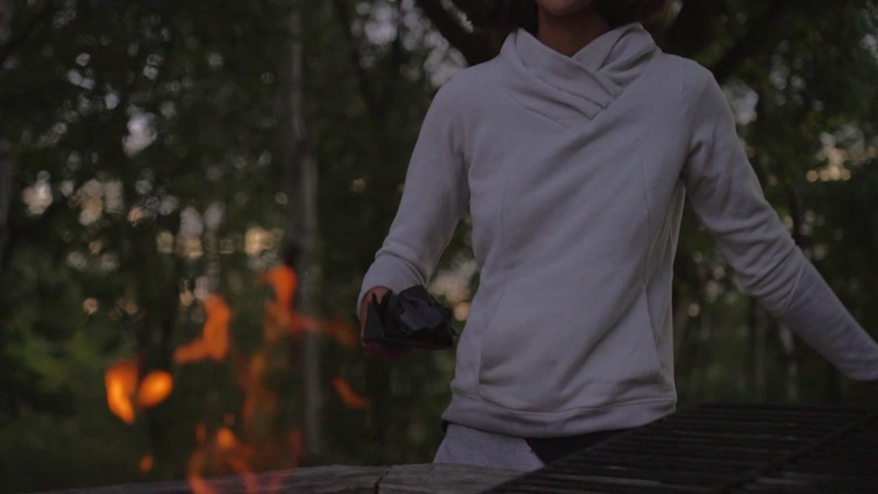 A woman with a joyful expression, putting coal into a fire outdoors at twilight