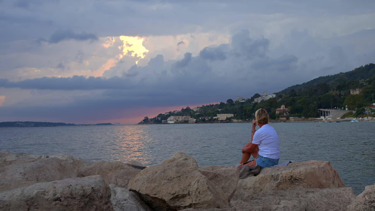 Blonde woman looking at the sea from rocks on the shore in the Golfe-Juan, France on a cloudy day at sunset