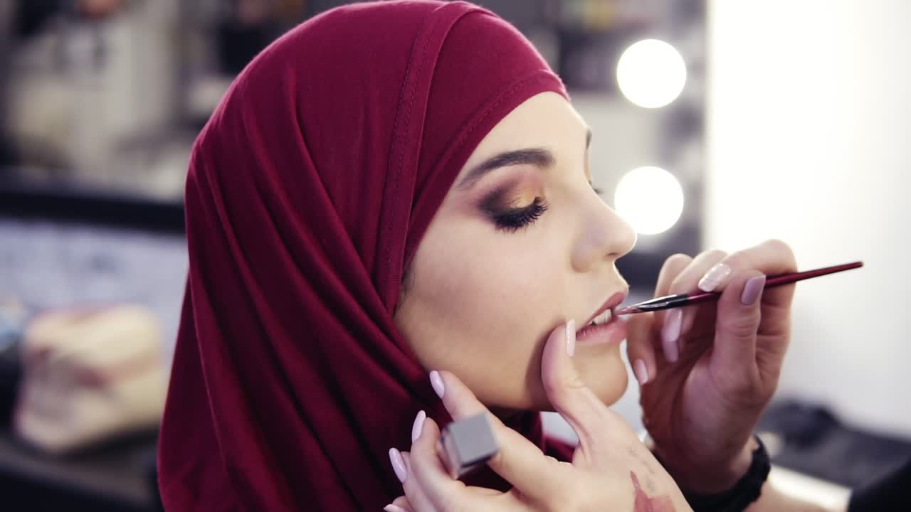 Unbelievably beautiful girl with hazelnut eyes and purple hijab on her head has unrecognizable make up artist's hand accurately applying lipstick with a special brush