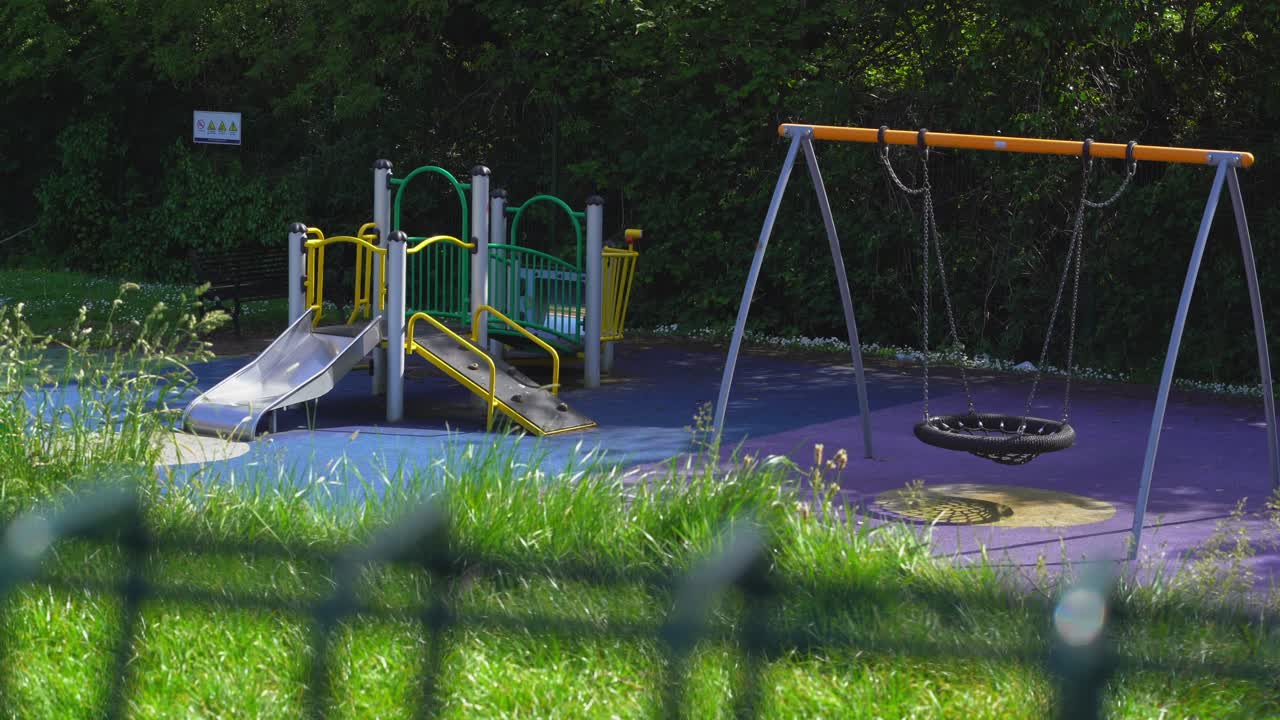 Empty playground through fence no children or kids green grass swing and climbing frame.