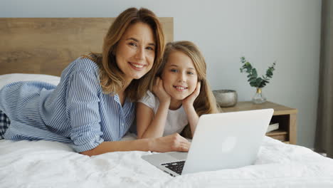 Joven-Y-Hermosa-Madre-Acostada-En-La-Cama-Con-Su-Hija-Frente-A-La-Laptop-Mientras-Sonríe-A-La-Cámara-Por-La-Mañana