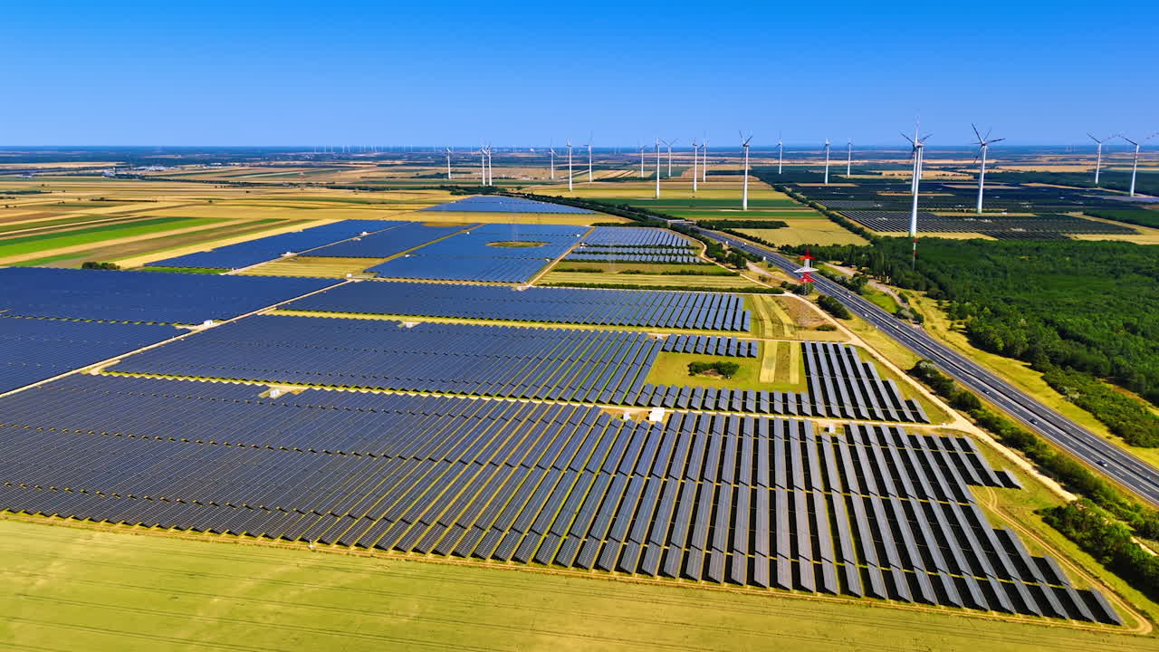 Multiple solar panels installed in the vast field. A highway goes along the site. Wind farm at backdrop. Aerial view