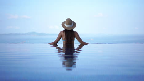 Reflejo-De-Espalda-Y-Agua-De-Mujer-En-Traje-De-Baño-Y-Sombrero-De-Pie-En-El-Borde-De-La-Piscina-Infinita-Y-Mirando-El-Horizonte