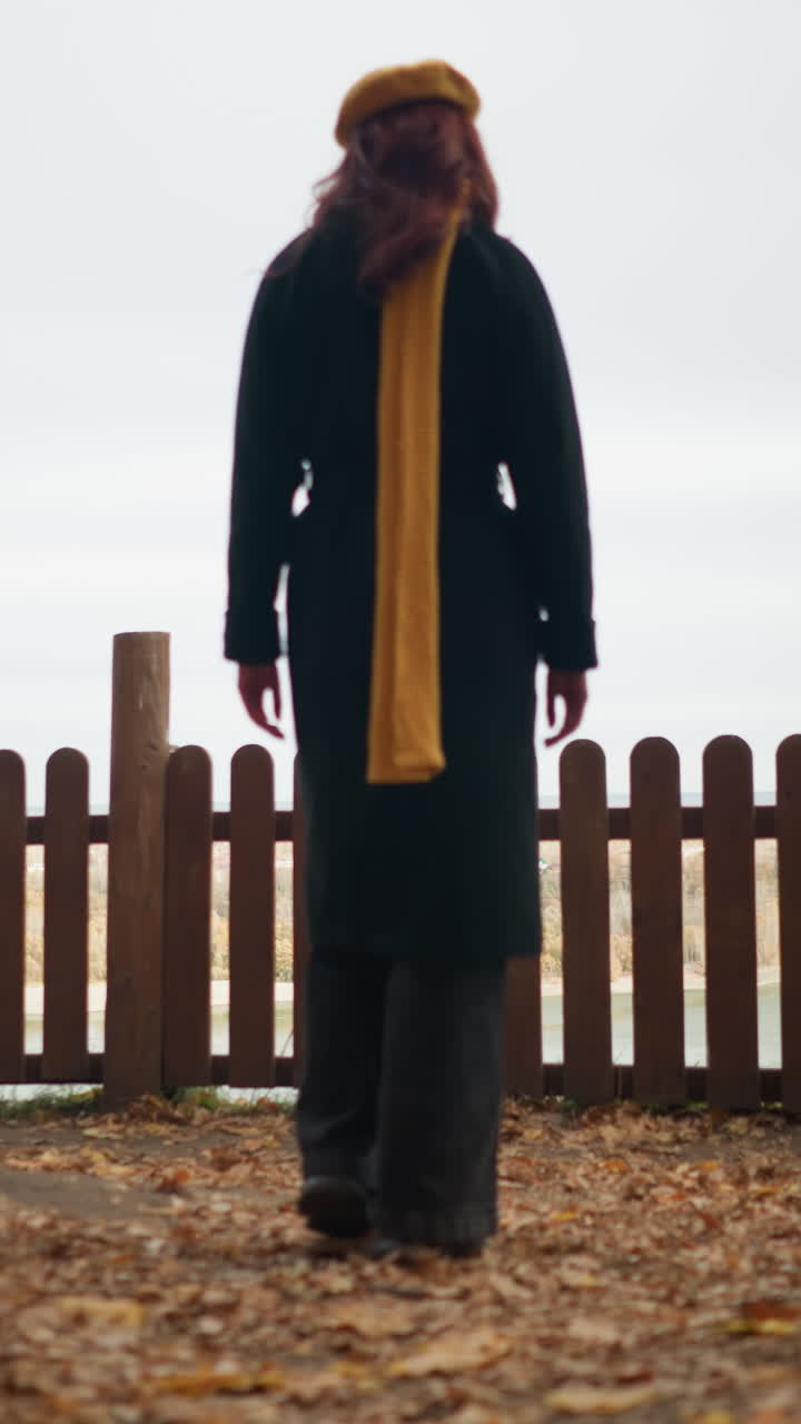 노란색 베레트와 검은색 코트를 입은 여자가 나무 울타리를 향해 걸어가며, 그 위에 손을 고, 건조한 가을 잎이 어져있는 울타리를 통해 강의 전망을 감상합니다.