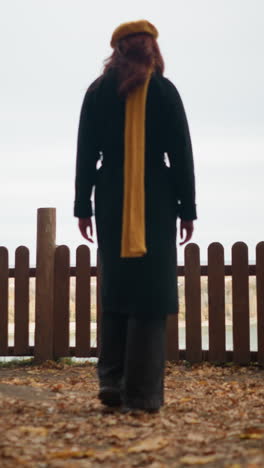 back view of a lady in a yellow beret and black coat walking toward a wooden fence, resting her hand on it, admiring the river view through the fence with dry autumn leaves scattered around