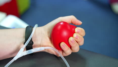 Female hand with black wristwatch or fitness bracelet is pressing red rubber heart. Tubes of a catheter are near the donor's hand. Blood donation concept. Close up.