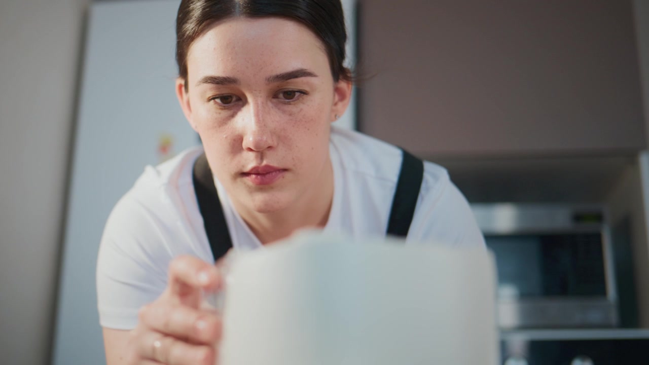Giovane donna in cucina, concentrata, che tiene una tazza. Appare seria e concentrata. Adatta per temi di caffè, colazione o lavoro. Illuminazione soffusa, interni moderni. Perfetta per lifestyle, cibo.