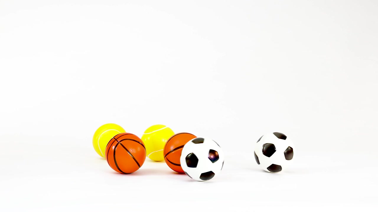 Small toy soccer, basketball, and tennis balls fall and bounce against a white studio background
