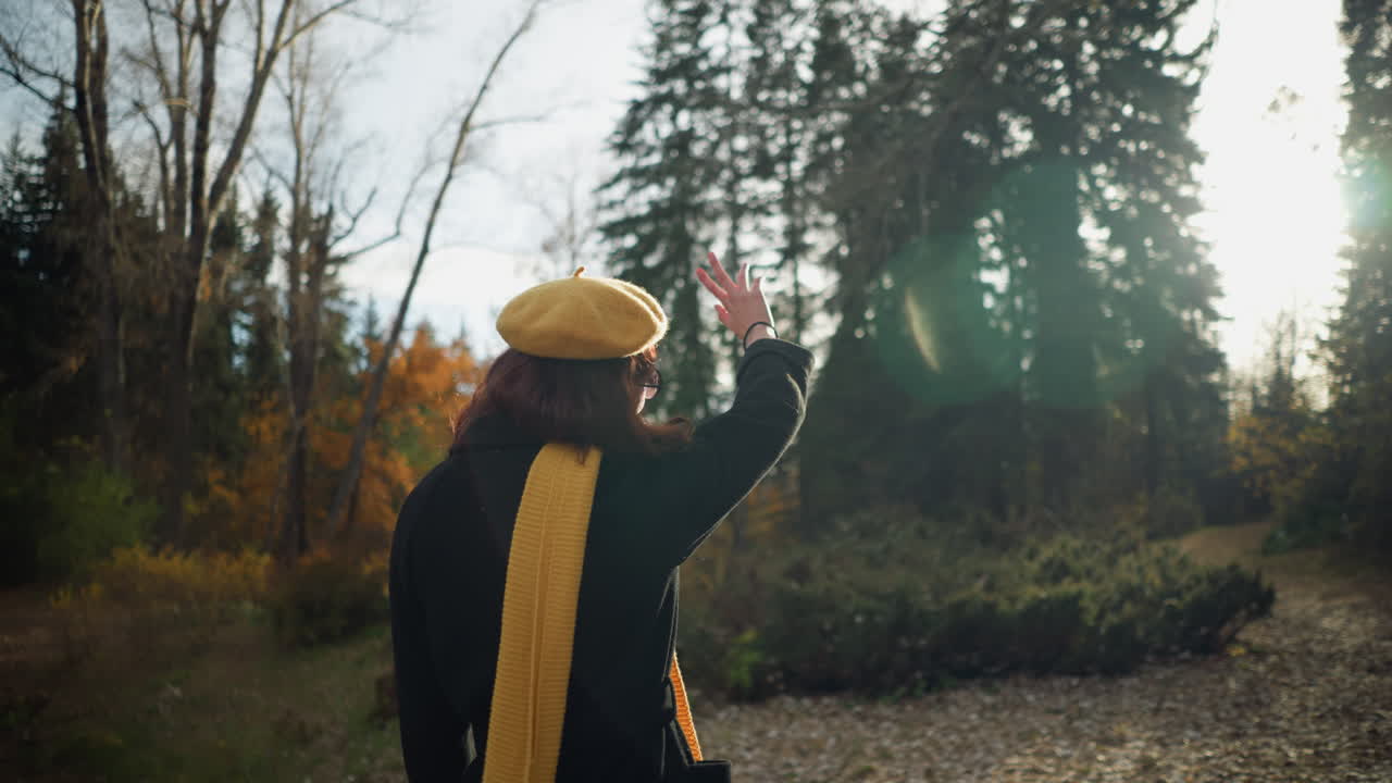 베레트와 머플러를 입은 여인이 숲길을 따라 걸어가며 손으로 빛을 가리키며, 빛은 그녀를 둘러싸고 따뜻한 빛을 만니다. 나무와 황금 잎자루로 둘러싸인 평화로운 가을 풍경을 즐기면서.