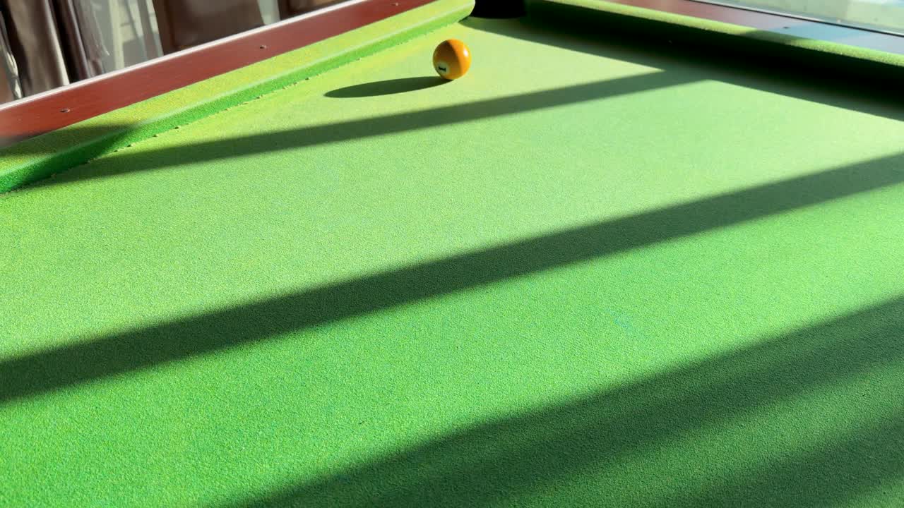 A white cue ball rolls across a sunlit green pool table, sinking a yellow ball