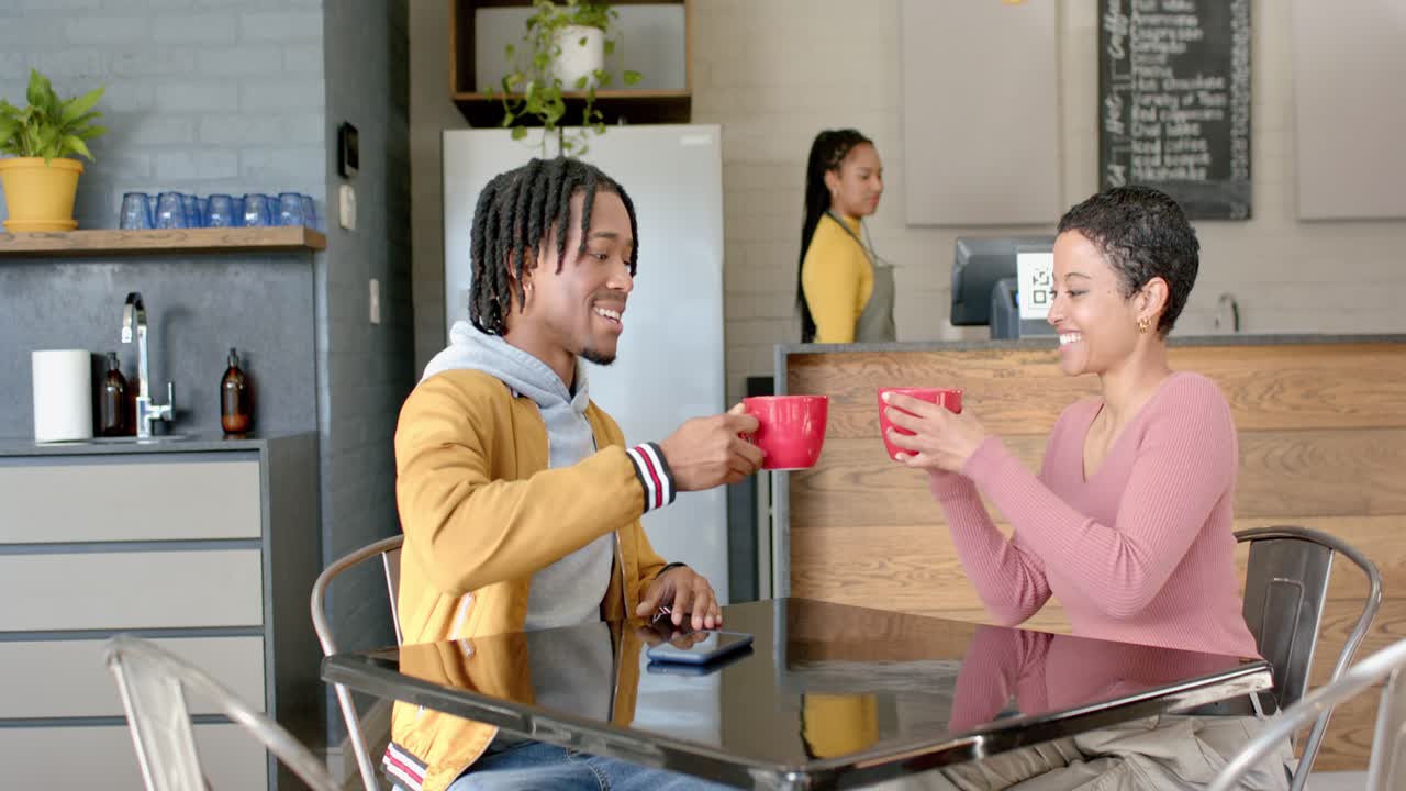 Diverse couple clinking red mugs on cafe table toasting date while barista operating register