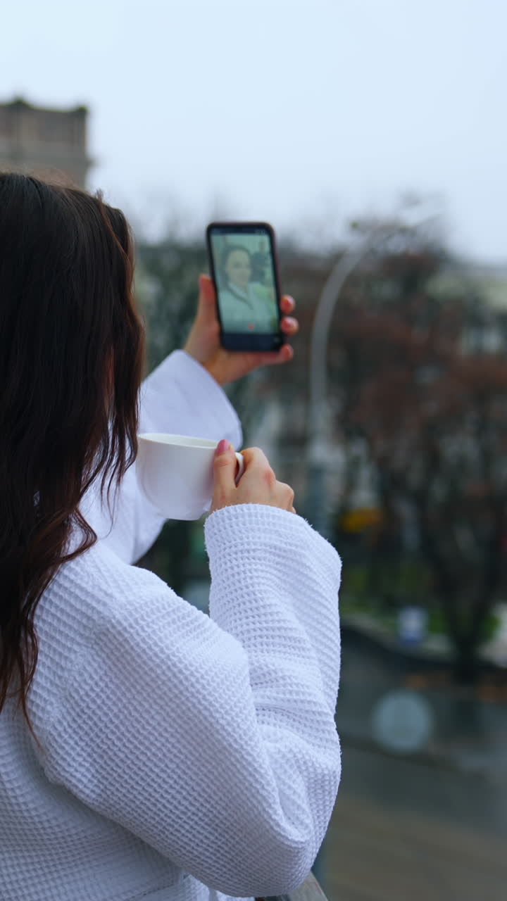 Young woman drinks coffee on a hotel balcony. Happy girl in a bathrobe doing selfie on the terrace on the urban background. Woman in white coat with a phone and a cup. Vertical video