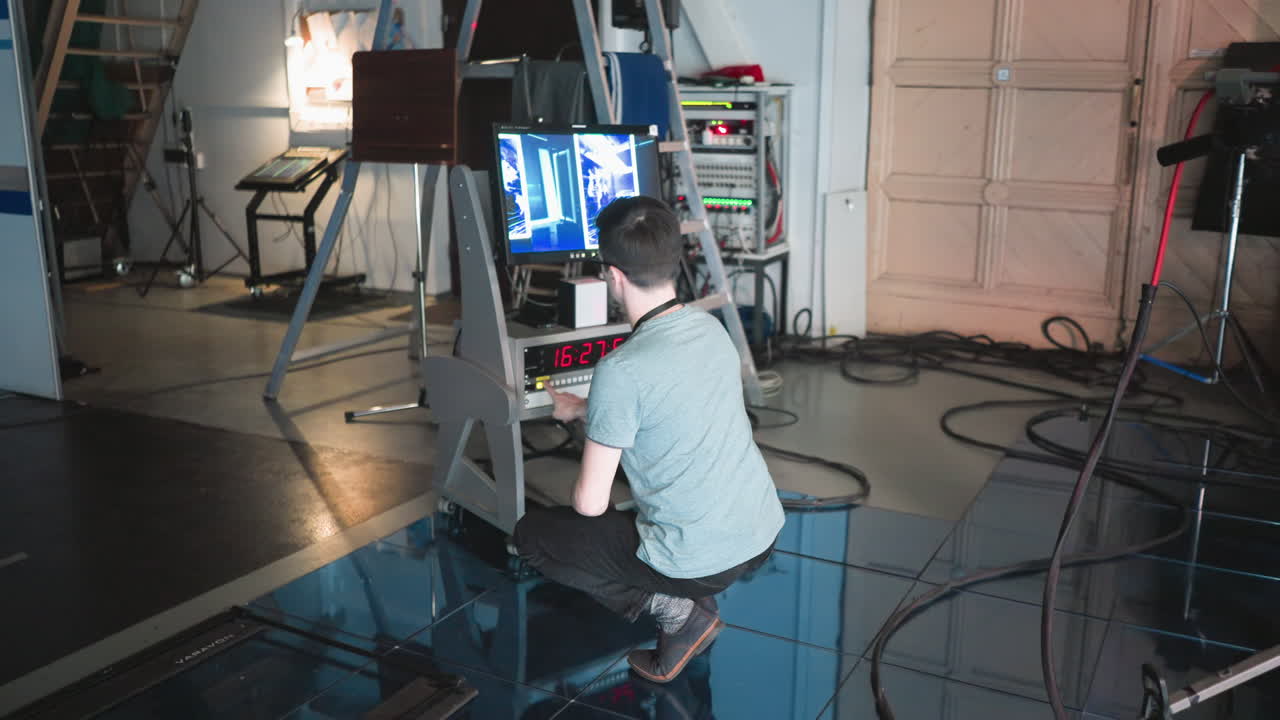Man crouching in professional studio, adjusting digital monitor and timer settings, focused on equipment in action, production underway, tech workspace with cameras and lights in background