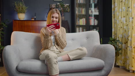 Mujer-Joven-Feliz-Usando-Teléfono-Inteligente-Y-Riendo-Mientras-Se-Sienta-En-Un-Sofá-En-Casa