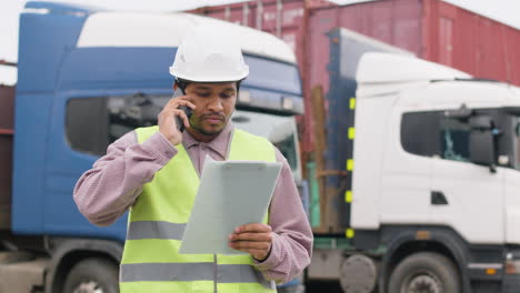 worker wearing vest and safety helmet organizing a truck fleet in a logistics park while consulting a document and talking on the phone 1