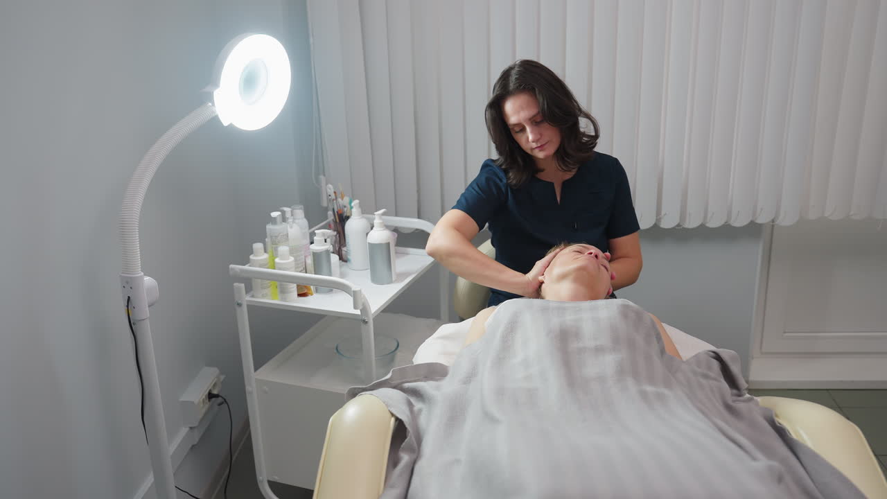 Beauty technician gives head and neck massage to client lying relaxed in spa treatment room with soft lighting and skincare products nearby for pampering and tension relief, client closes eyes
