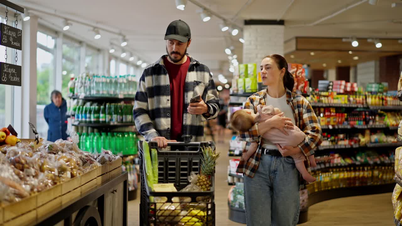A confident man in a cap along with his wife and small infant child walk through the supermarket and choose the necessary products during their shopping