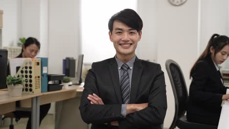 portrait of a successful asian male team leader wearing business suit looking at camera with smile and folded arms at modern workplace background with coworkers