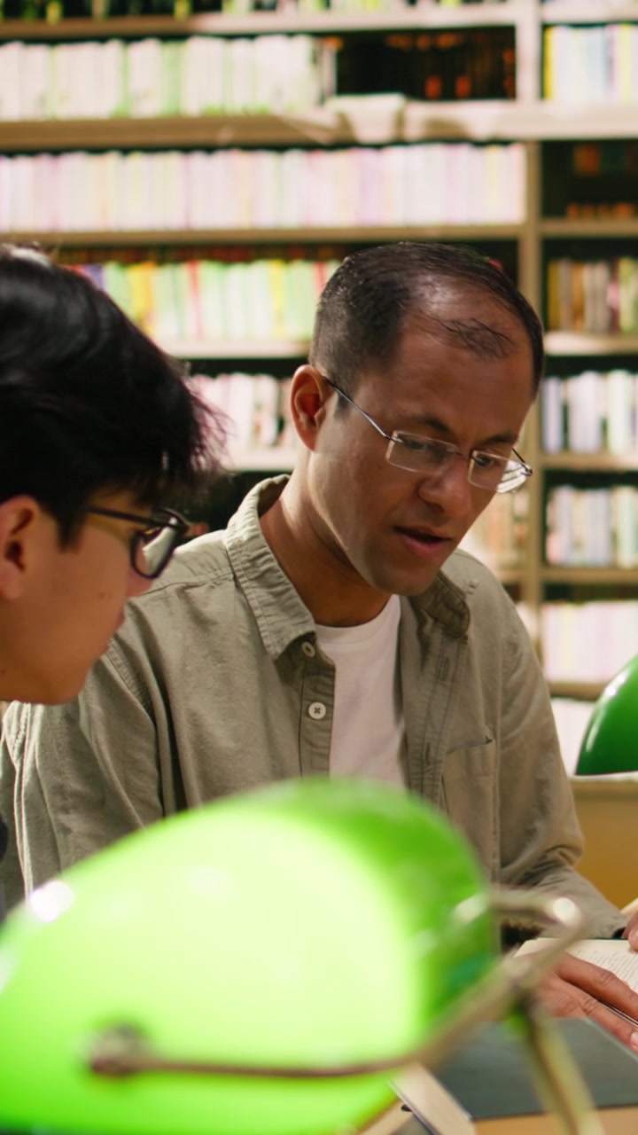 Video Vertikal Mentor dan mahasiswa mendiskusikan penelitian di atas buku-buku terbuka di meja perpustakaan kayu