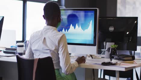 afrikanisch-amerikanische frau mit gesichtsmaske sitzt auf ihrem schreibtisch in einem modernen büro und benutzt einen computer