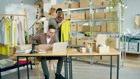 african american designer woman typing on tablet and checking boxes while talking with female designer coworker in fashion clothing shop