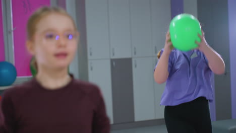 young girls in gym working out, juggling and throwing green balloon up with large exercise ball and light shining in background, demonstrating coordination and fitness activities