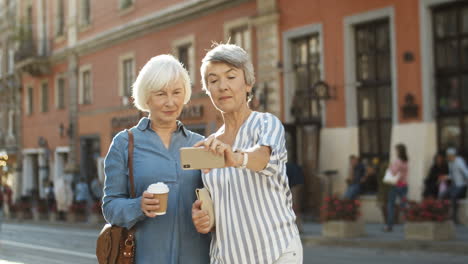 Two-Beautiful-Old-Women-Smiling-While-Taking-Selfie-Photo-With-Smartphone-Camera-On-Street-In-City-Center