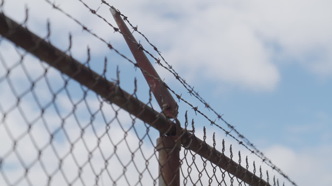 Rusty barbed wire fence in private property or closed-off area in backyard or prison. Close up on sharp wire against intruders or trespassing. No entry. Clouds in sky. Scrape-yard in rural place