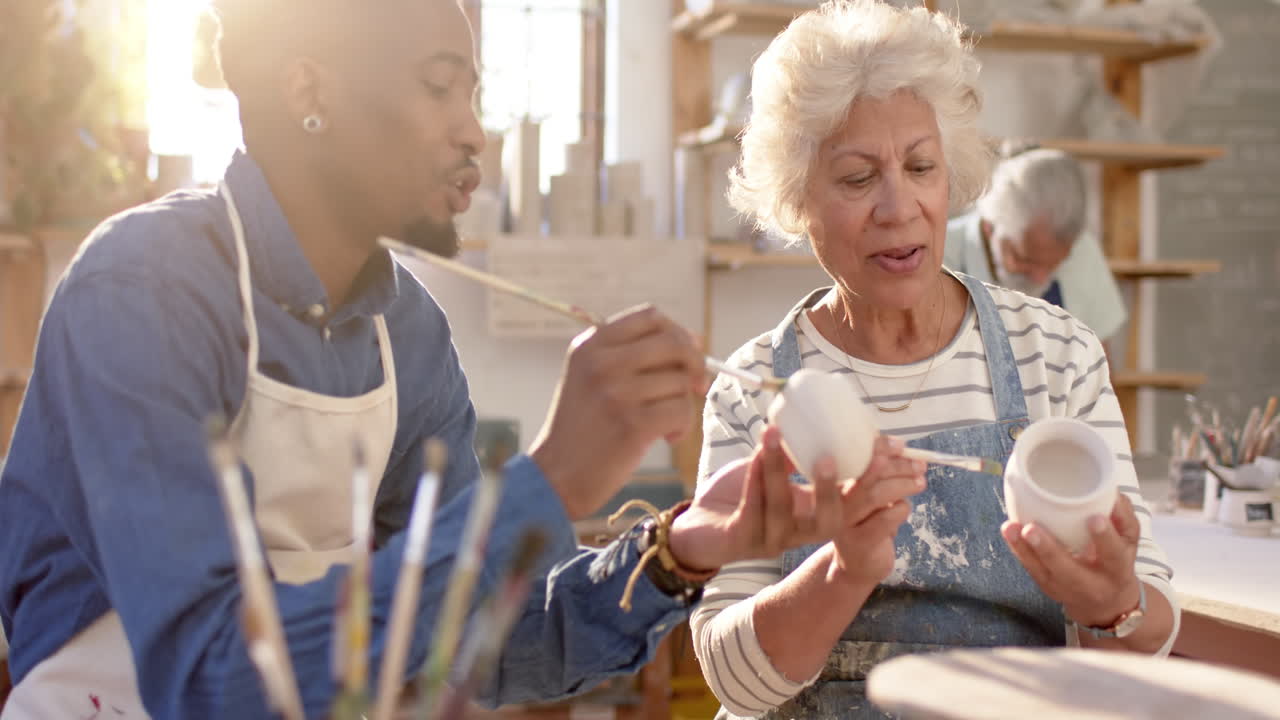 Two diverse male and female potters glazing clay jug and discussing in pottery studio, slow motion