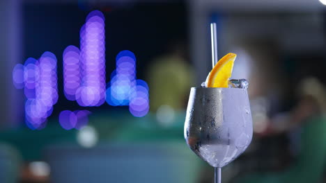 rotating metal wineglass with misted surface. cocktail with ice and orange slice on top and metal straw inside. close up. blurred backdrop.