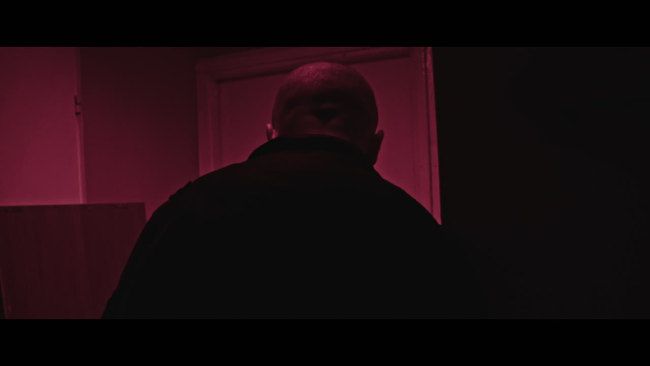 A bald dangerous looking man stands in the red corridor