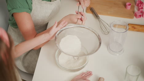little girl taps sieve with left hand while instructor holds handle steady, flour falling into glass bowl, baking utensils scattered on white table, partial view of another child playing in background