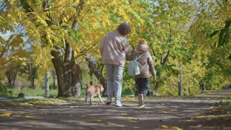 woman holds dog leash as it pulls ahead while daughter holds mother hand and looks up into golden autumn canopy, leaves gently falling on path, warm family moment of playful stroll under trees