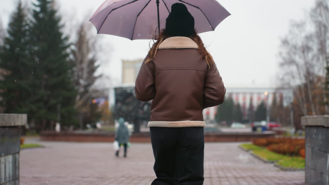 Back view of female walking in light snowfall holding umbrella wearing knit cap brown shearling jacket black trousers one hand pocket surrounded by autumn leaves, serene outdoor atmosphere