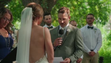 groom reads his vows from a paper to the bride while groomsmen stand behind him