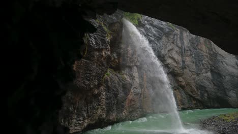 tall waterfall cascading into a turquoise river below, view from inside a rocky cave