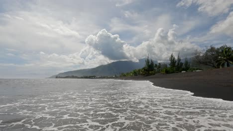 Playa-De-Taharuu-En-Tahití,-En-La-Polinesia-Francesa,-Con-Arena-Negra-Y-Olas-Del-Océano-Pacífico-Y-Surfistas-Al-Fondo